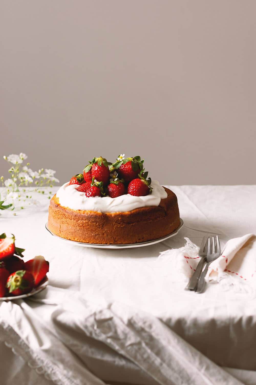 En una mesa hay un plato blanco y en el plato está el bizcocho de fresas y yogur decoradas con nata montada y fresas.
