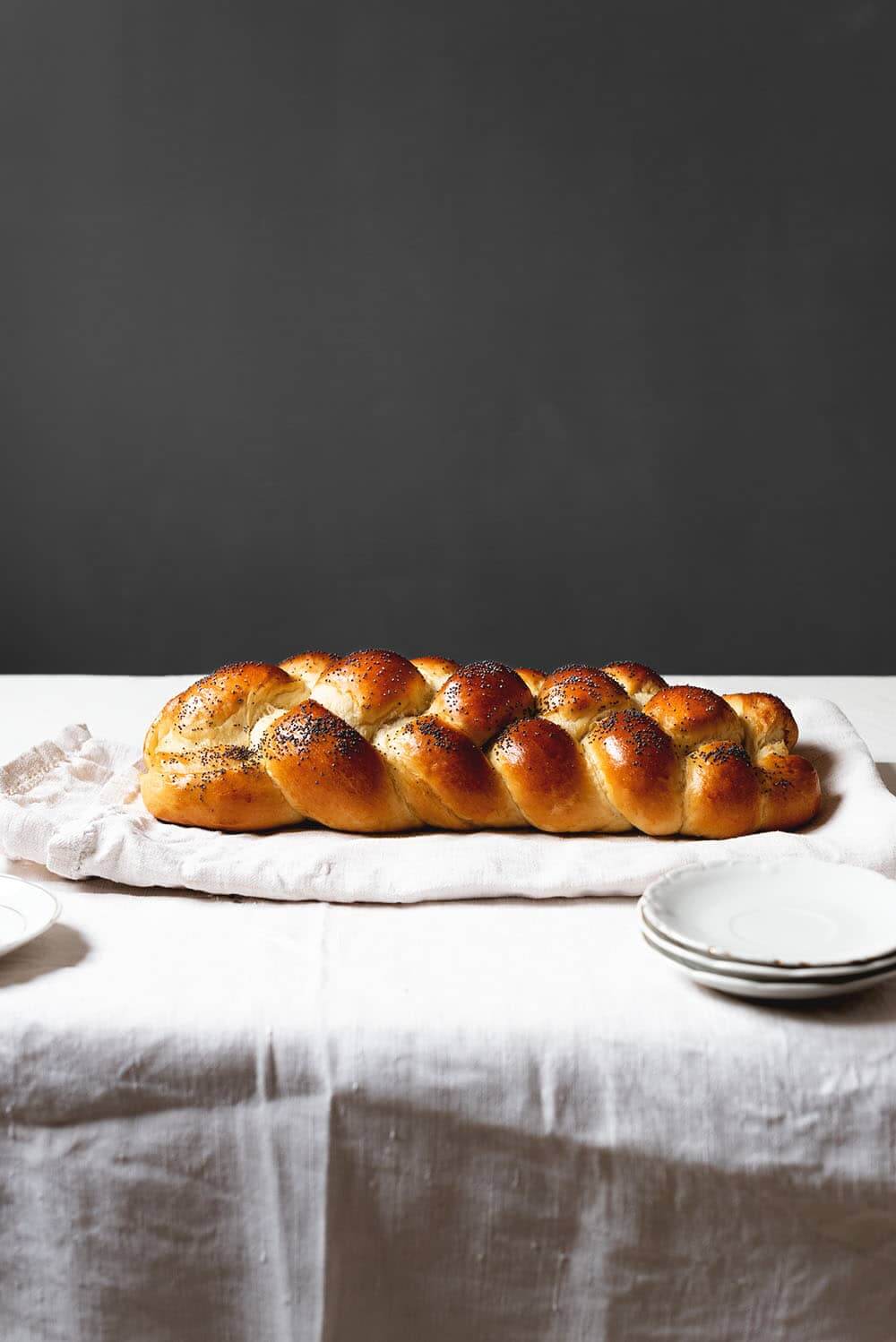 En un paño de cocina blanca está puesto el pan judio sin cortar. Se ve el trenzado de cuatro cabos bonito y la superficie del pan brillante. Delante del pan hay un par de platos blancos.