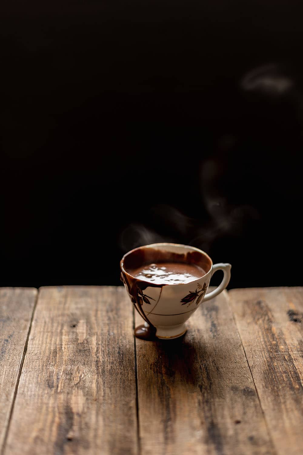 En una mesa de madera está una taza de chocolate caliente humeando.