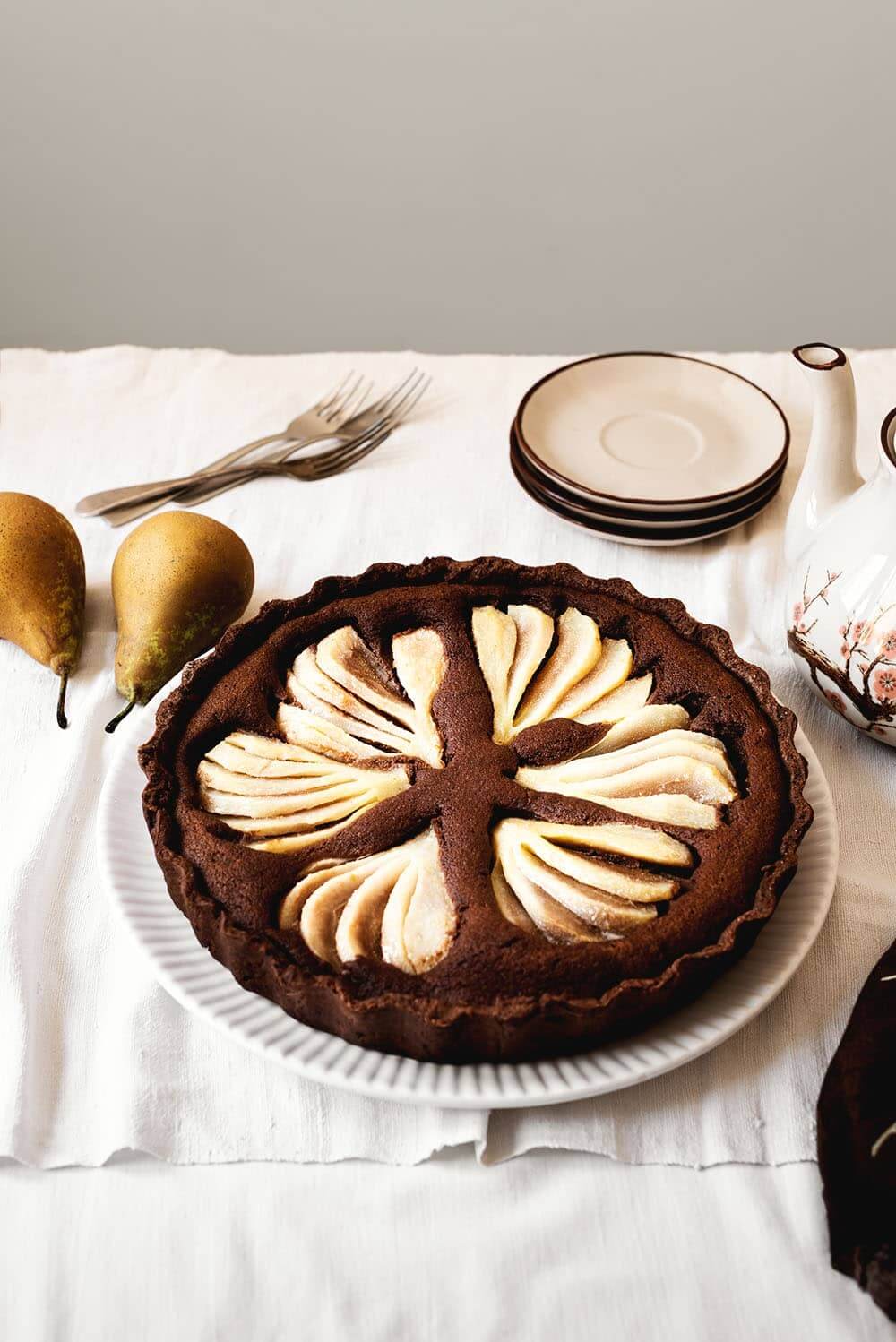 En una mesa está un mantel blanco. En una fuente grande y blanca está la tarta de peras con chocolate. En la mesa hay una tetera, platos pequeños, cubiertos también.