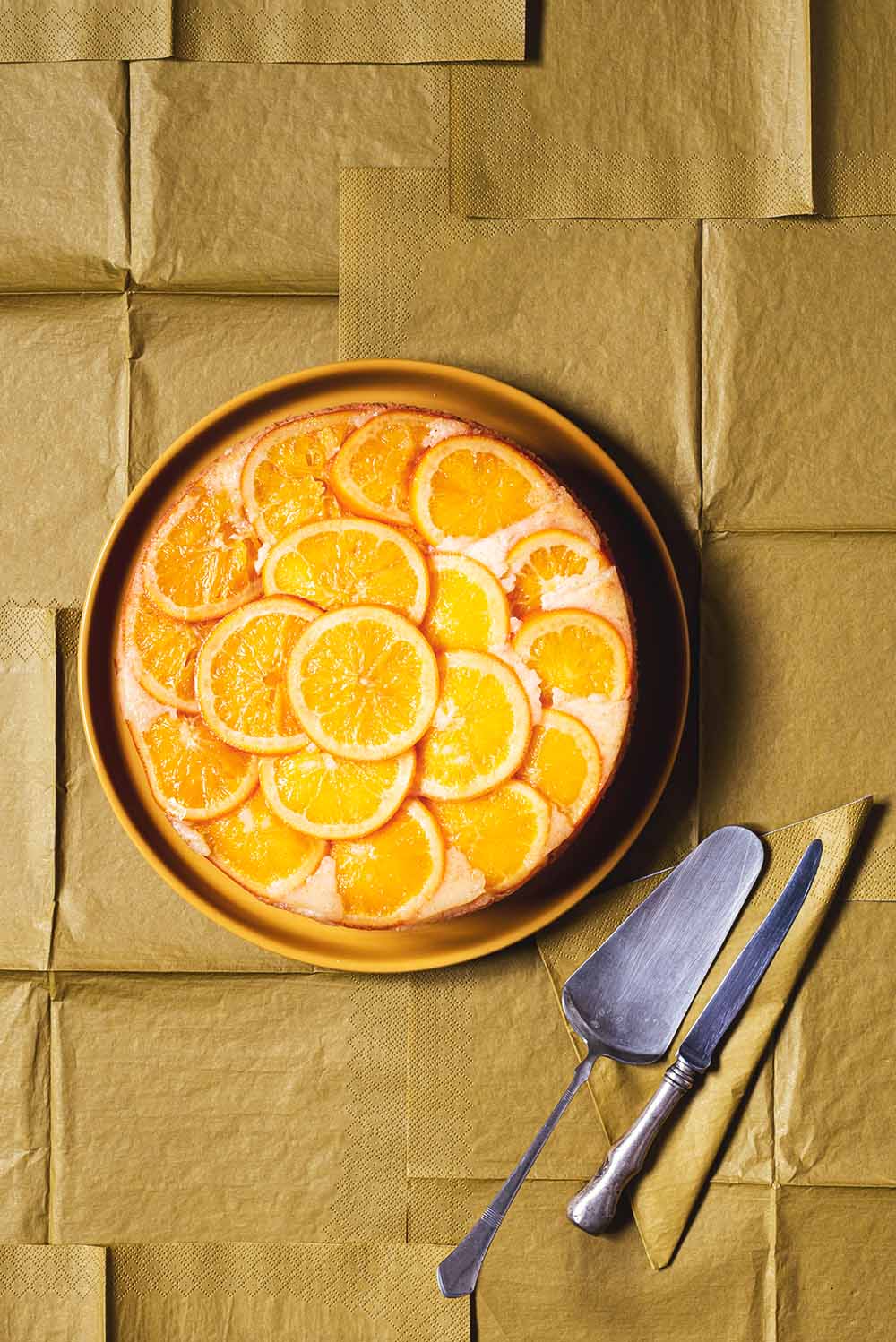 En un mantel de color caqui está puesto un plato de color naranja y encima de este plato está la tarta de naranja invertida. También en el plato hay un cuchillo para cortar.