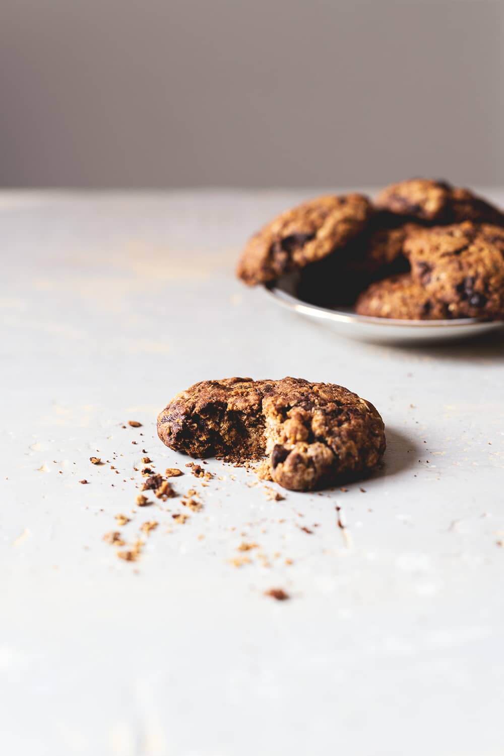 En una superficie gris está una galleta de galleta de avena mordida. Detrás se ve en un plato muchas galletas de avena y chocolate amontonados.
