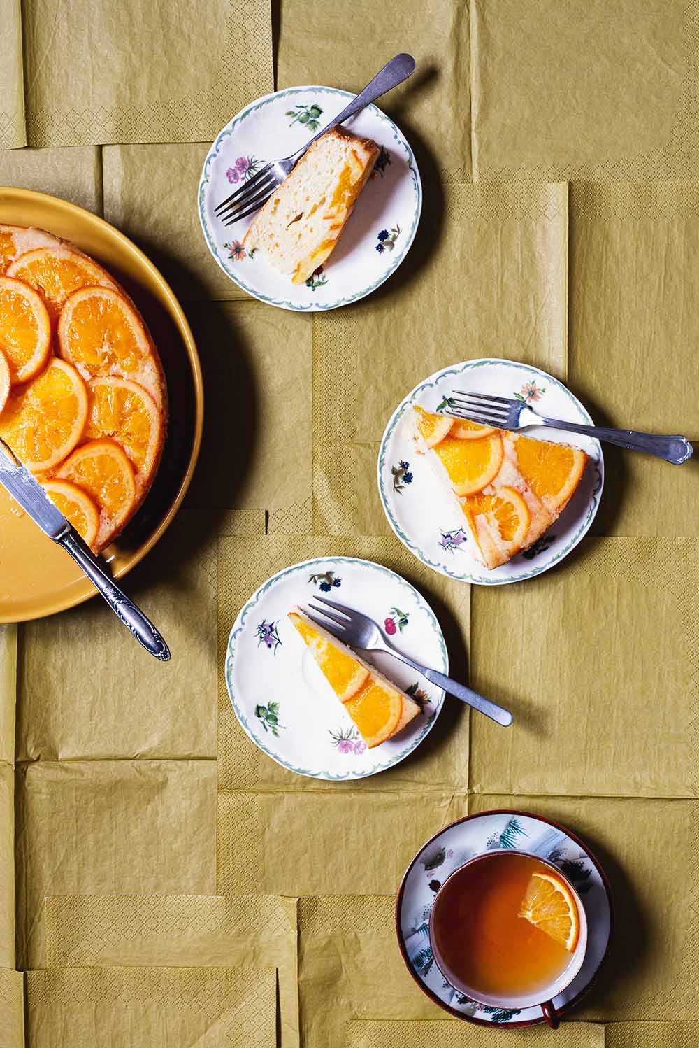 En un mantel de color caqui está la tarta de naranja invertida ya cortada. En dos platos pequeños hay dos raciones de tarta. En cada plato hay un tenedor. En el mantel también se ve una taza de té.