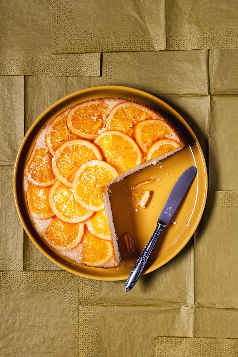 En un mantel de color caqui está puesto un plato de coclor naranja y encima este plato está la tarta de naranja invertida. También en el plato hay un cuchillo para cortar.
