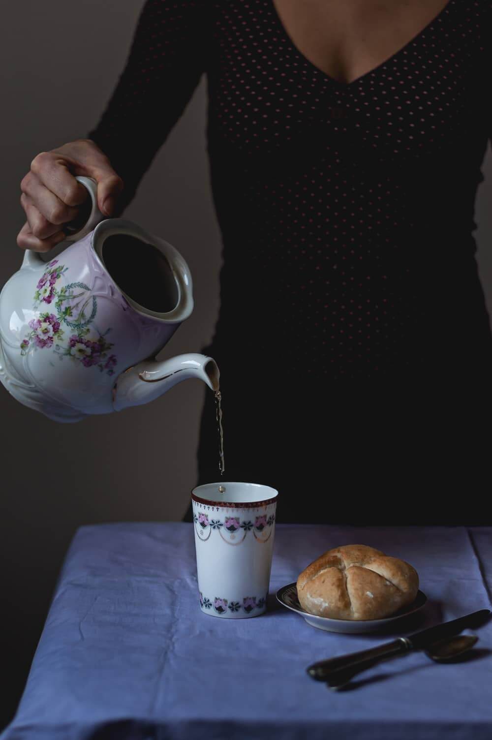 Una mesa esta cubierta con una mantel de color lila. Una persona está echando té recién hecho a una taza. Al lado de la taza en un plato pequeño está el Panecillo de Kaiser.