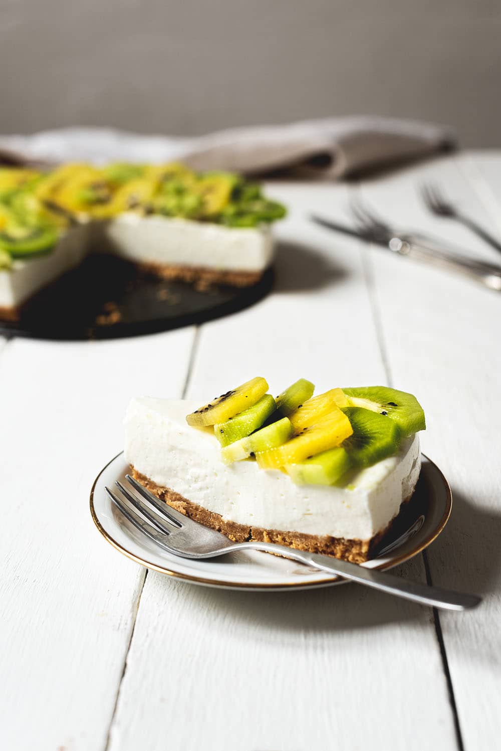 En una mesa de madera blanca está puesta una ración hermosa de tarta de kiwi y yogur. Detrás se ve la tarta, unos cubiertos y un paño de cocina.