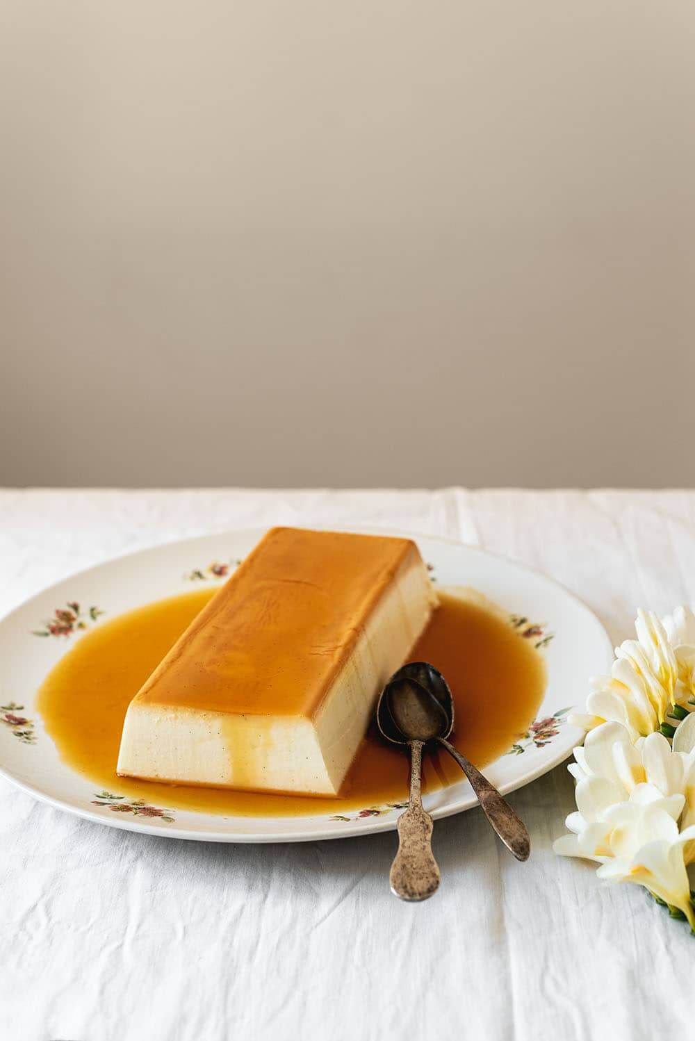 En una mesa cubierta con un mantel blanco hay un plato blanco con el flan de queso sin horno. El flan está rodeada con la salsa de caramelo. Al lado del plato hay unas flores blancas.
