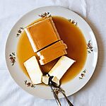En una mesa cubierta con un mantel blanco hay un plato blanco con el flan de queso sin horno. Ya está cortada y está rodeada con la salsa de caramelo.