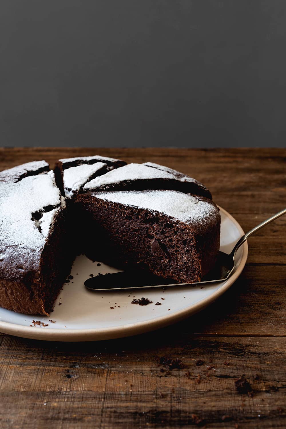 En una mesa de madera está el bizcocho de chocolate con aceite de oliva cortado y espolvoreado con azúcar glas. También se ve una pala para tartas.