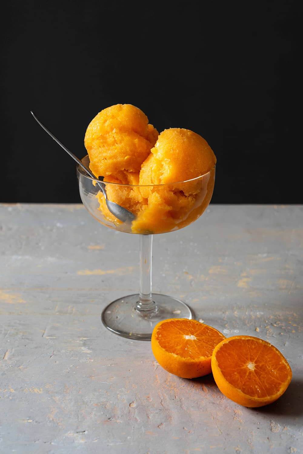 En una superficie gris está una copa de helado. Dentro de la copa hay varias bolas de sorbete de mandarina sin alcohol. Alrededor hay mandarinas cortadas en la mitad.