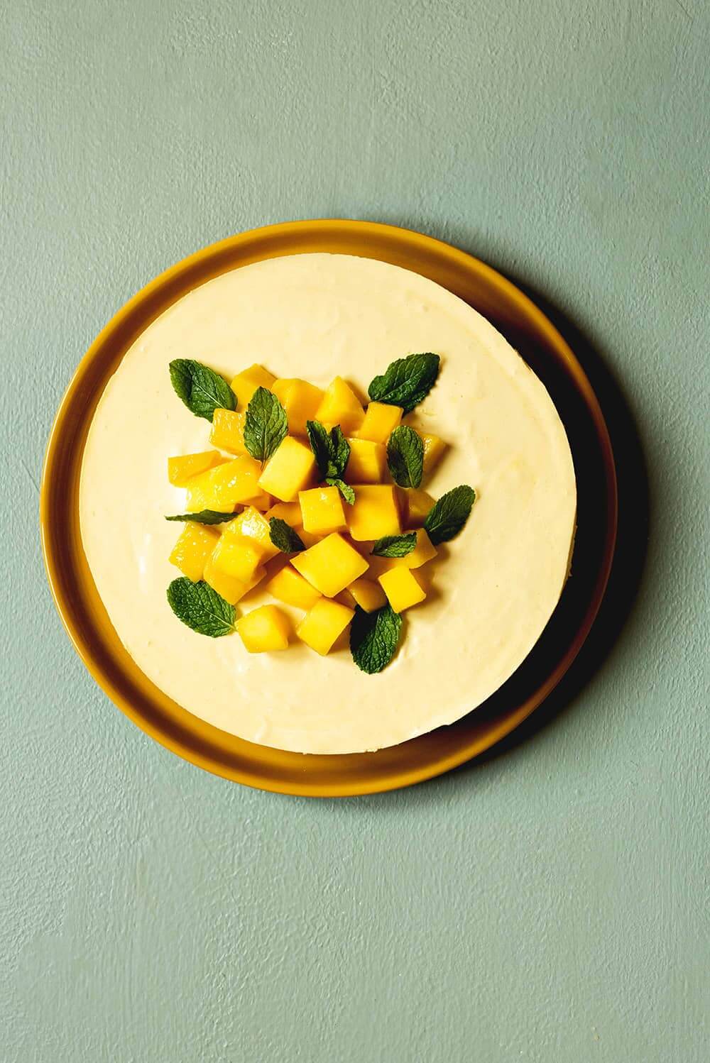 En una superficie verde, está un plato amarillo. En el plato tarta de mango y mascarpone. El postre está decorado con mangos frescos. La tarta está sin cortar. El postre está decorado con mangos frescos.