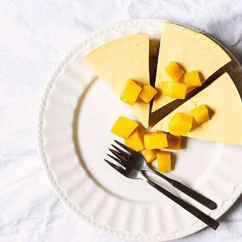 En una mesa cubierta con un mantel blanco, está un plato. En el plato blanco están tres trozos de tarta de mango con bizcocho. El postre está decorado con mangos frescos.