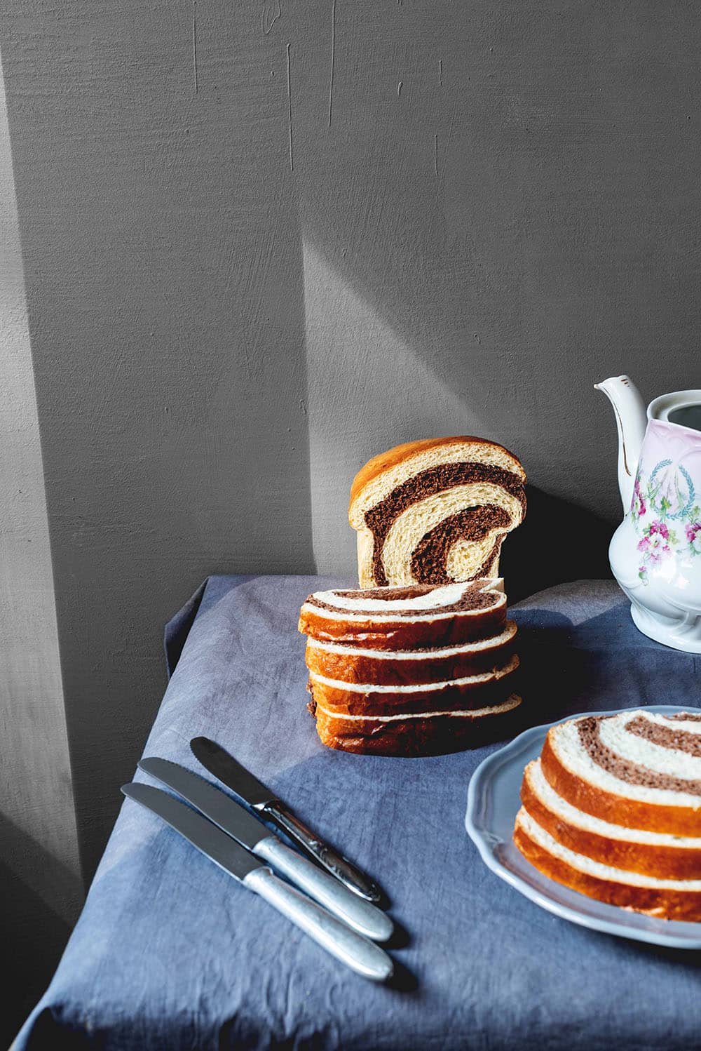 En una mesa cubierta con un mantel gris está el pan de brioche marmolado cortado. Alrededor hay una tetera vintage y en un plato hay más pan dulce marmolado cortado.