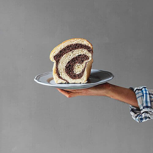 Delante de una pared gris una persona está sujetando un plato. En este plato está un trozo de pan de brioche marmolado cortado.