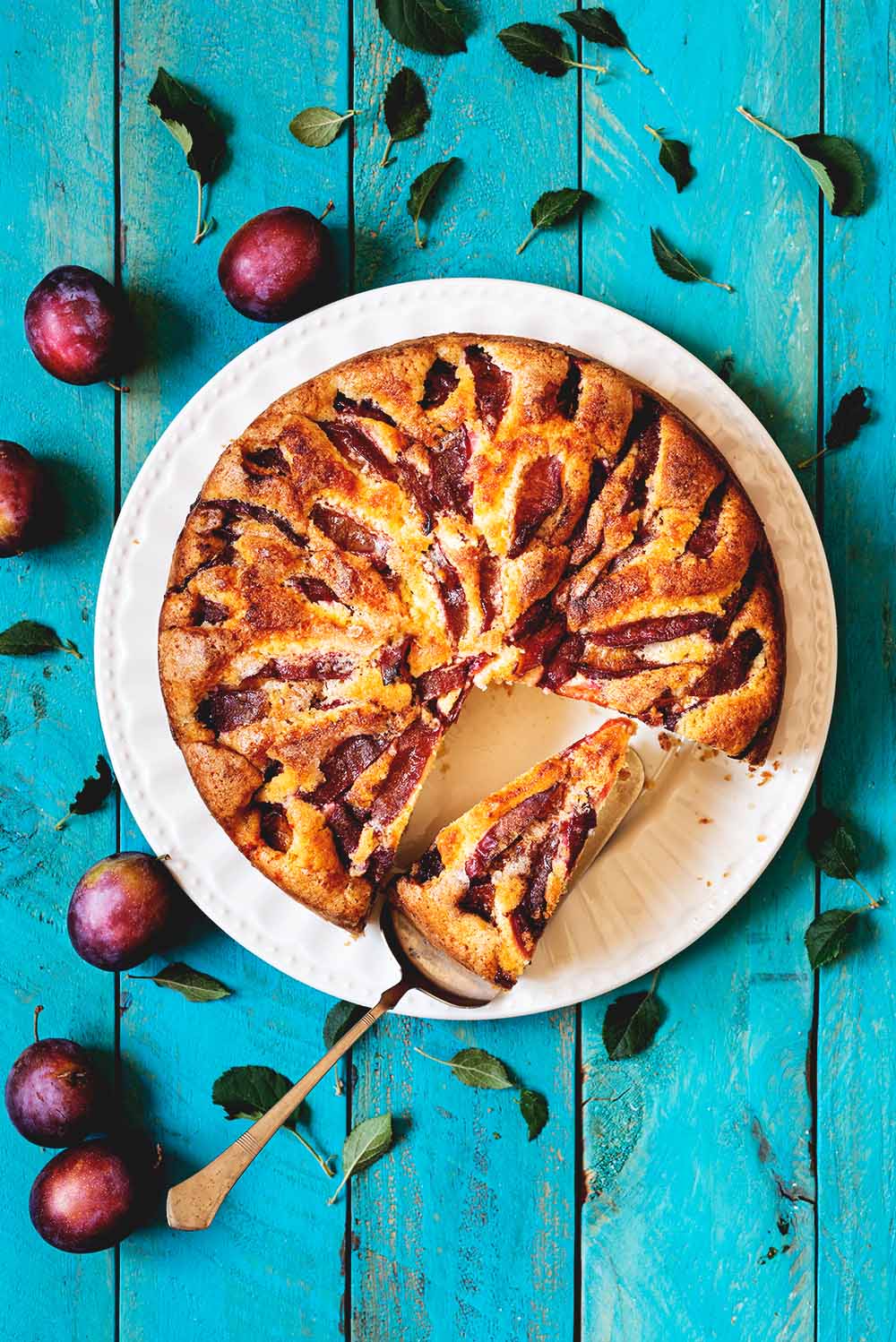 En una mesa azul está el bizcocho de ciruelas rojas y canela. Al lado del plato hay ciruelas rojas con hojas. En el plato hay una pala de tarta con una ración de bizcocho.