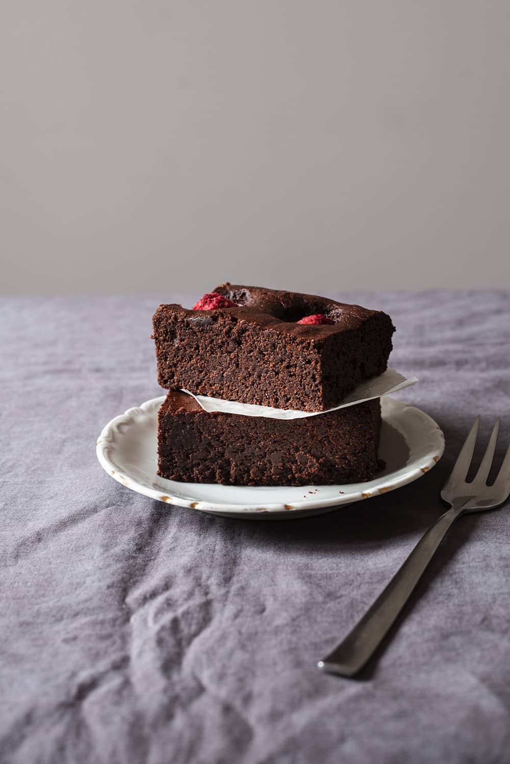 En una mesa cubierta con un mantel gris hay un plato blanco. Encima hay dos raciones de brownie de frambuesas formando un torre. Entre las dos raciones hay un trozo de papel de horno. Se ve muy jugoso. Al lado del plato hay un tenedor.