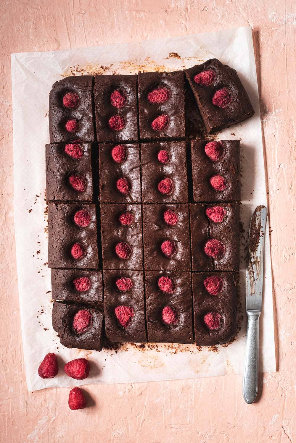 En una mesa rosa está un papel de horno. Encima está el brownie de frambuesas. Ya está cortado formas rectangulares. Se ve muy jugoso. Al lado del bizcocho hay frambuesas frescas y un cuchillo. 