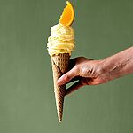 Delante de una pared verde una persona está sujetando un cucurucho lleno de helado de naranja casero. Como decoración hay un gajo de naranja encima de última bola de helado. 