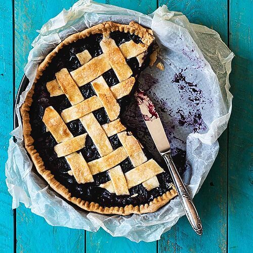 En una mesa azul está el pie de arándanos en su molde. Está cortado y se ve un cuchillo que está apoyando en la pared del mole.