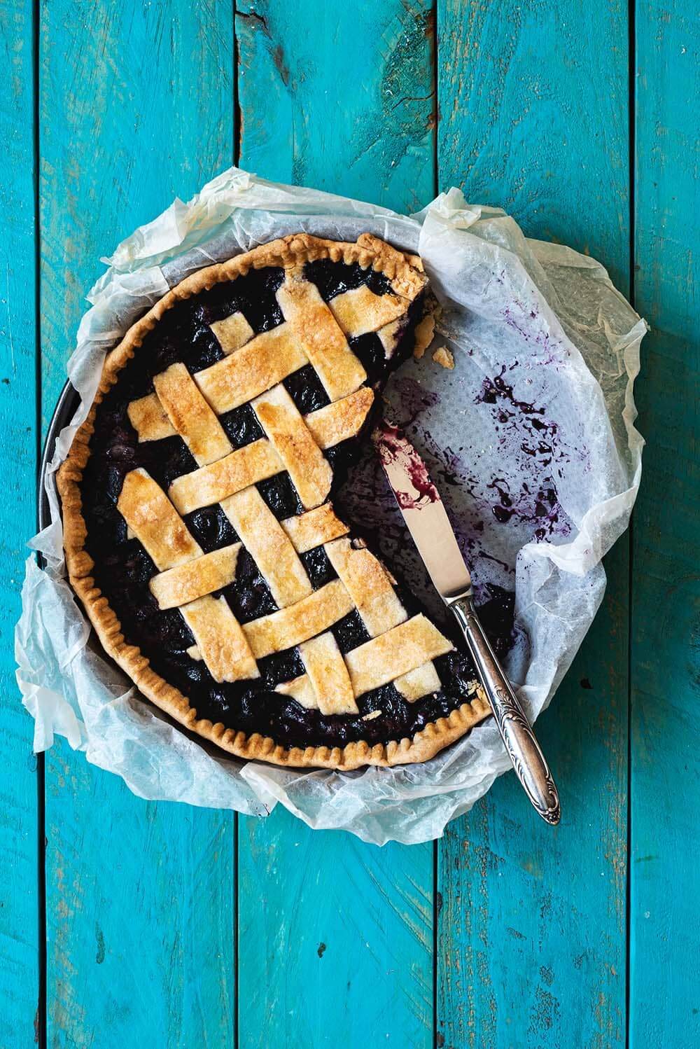 En una mesa azul está el pie de arándanos en su molde. Está cortado y se ve un cuchillo que está apoyando en la pared del mole.