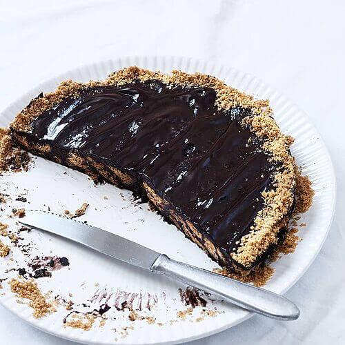 En una mesa cubierta con mantel blanco hay un plato blanco. Encima está la tarta de chocolate con galleta ya cortada. Al lado del postre hay un cuchillo.