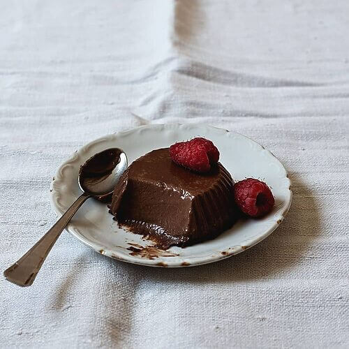 En una mesa cubierta con mantel blanco hay un plato pequeño. Encima está la panna cotta de chocolate. Le falta un bocado y está decorada con una frambuesa. Al lado hay una cuchara pequeña.