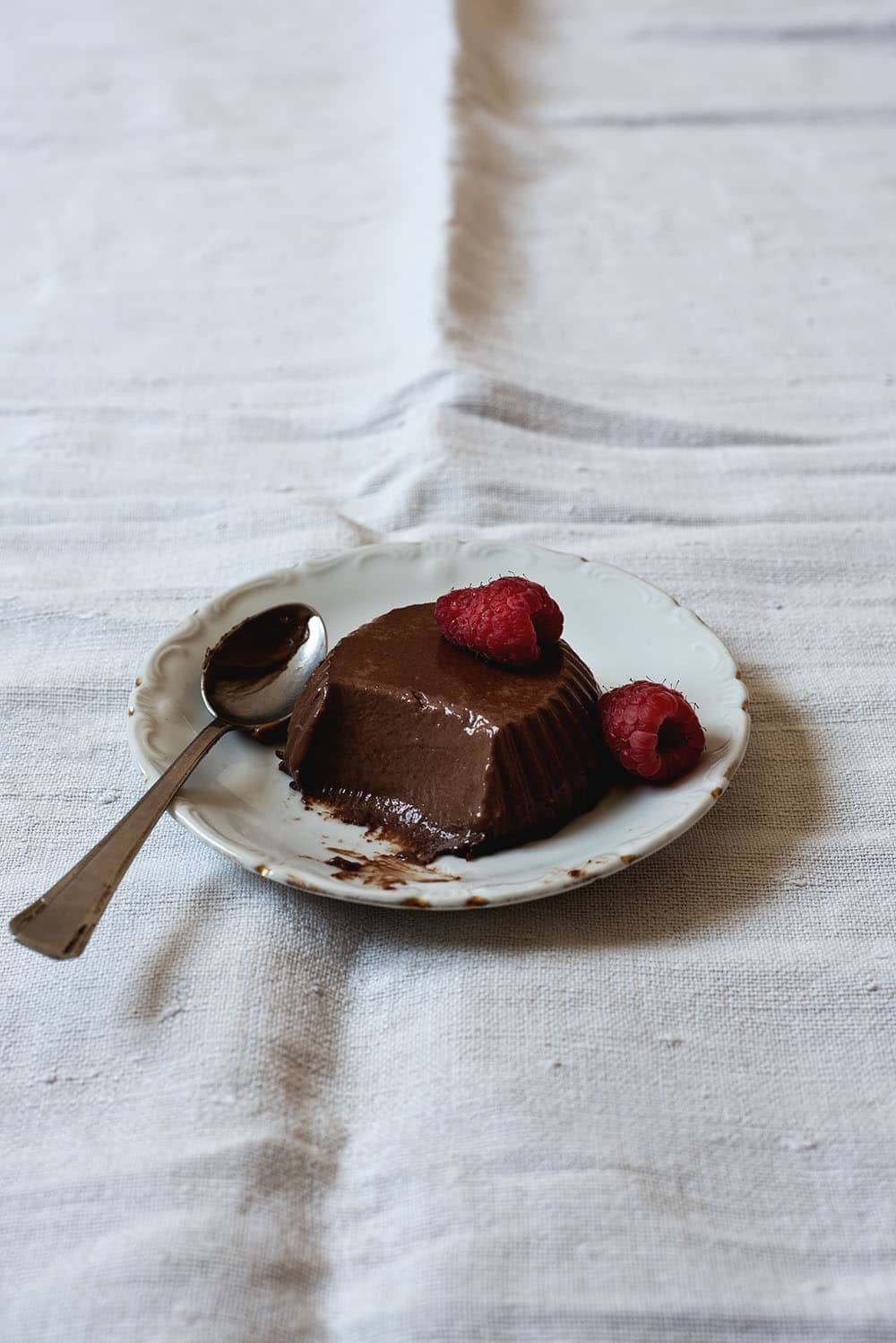 En una mesa cubierta con mantel blanco hay un plato pequeño. Encima está la panna cotta de chocolate. Le falta un bocado y está decorada con una frambuesa. Al lado hay una cuchara pequeña.