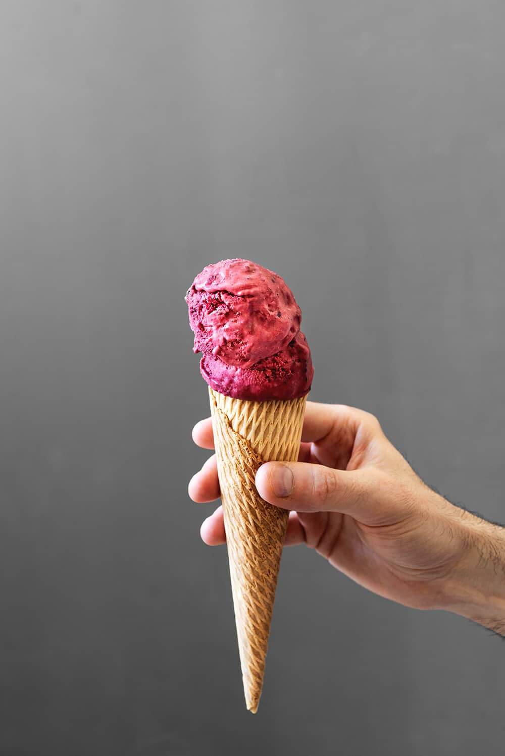 Delante de una pared gris una persona está sujetando un cucurucho. En él hay varias bolas de helado casero de mora. Se ve muy cremoso.