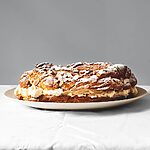 En una mesa cubierta con un mantel blanco hay un plato blanco. Encima está el Paris Brest. Está espolvoreado con azúcar glas.