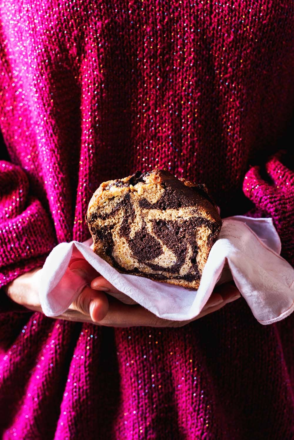 Una pesona vestida con un jersey de color rosa, esta sujetando en la mano un trozo de bizcocho de plátano marmolado que está envuelto con un paño de cocina rosa. Se ve muy esponjoso.