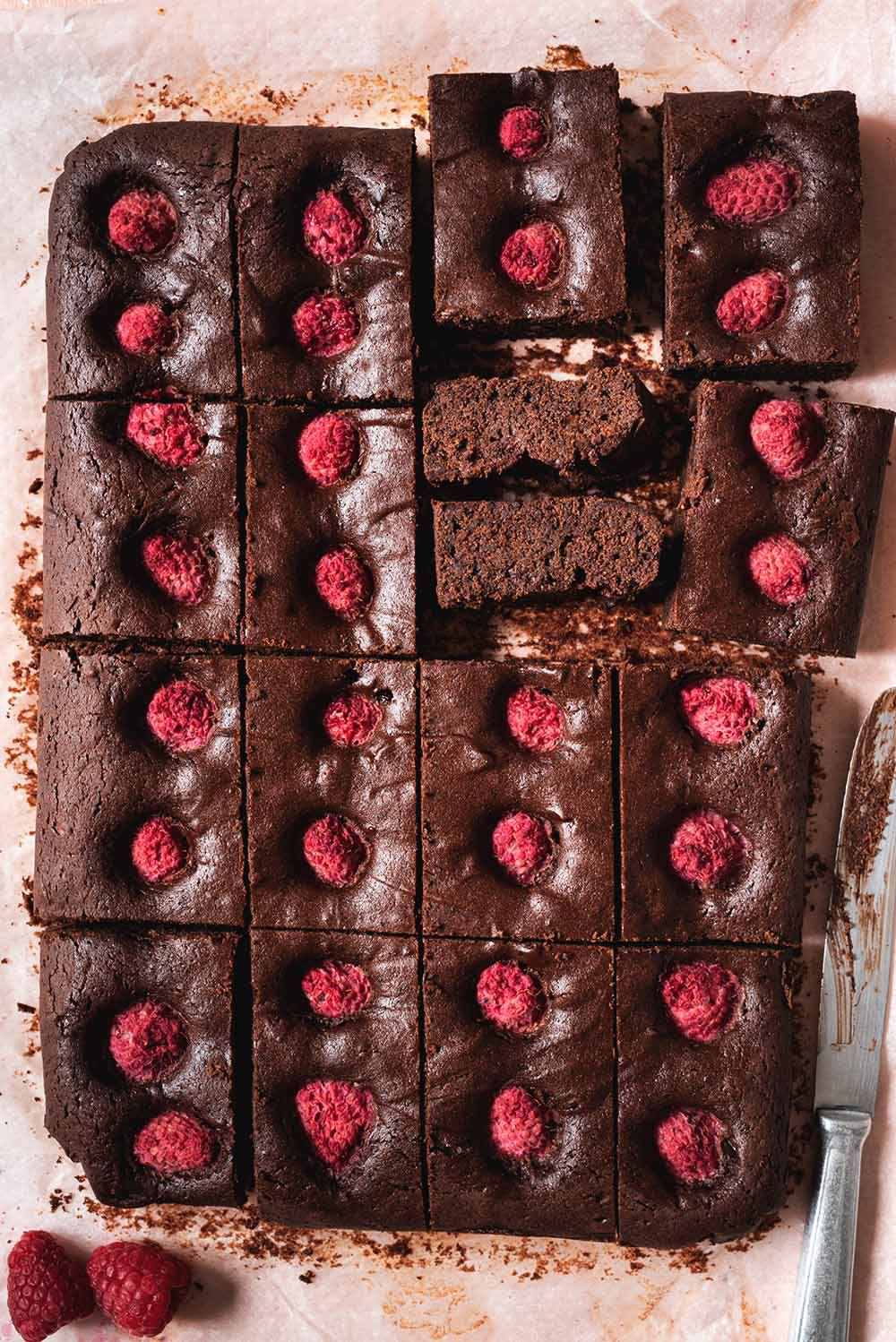 En una mesa rosa está un papel de horno. Encima está el brownie de frambuesas. Ya está cortado formas rectangulares. Hay dos trozos que enseña en interior. Se ve muy jugoso. Al lado del bizcocho hay frambuesas frescas y un cuchillo.