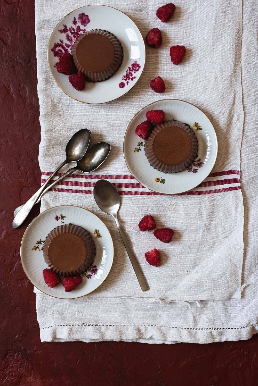 En una mesa burdeos cubierta con mantel blanco hay tres platos pequeños. Encima de cada una está la panna cotta de chocolate. Alrededor hay cucharas pequeñas.