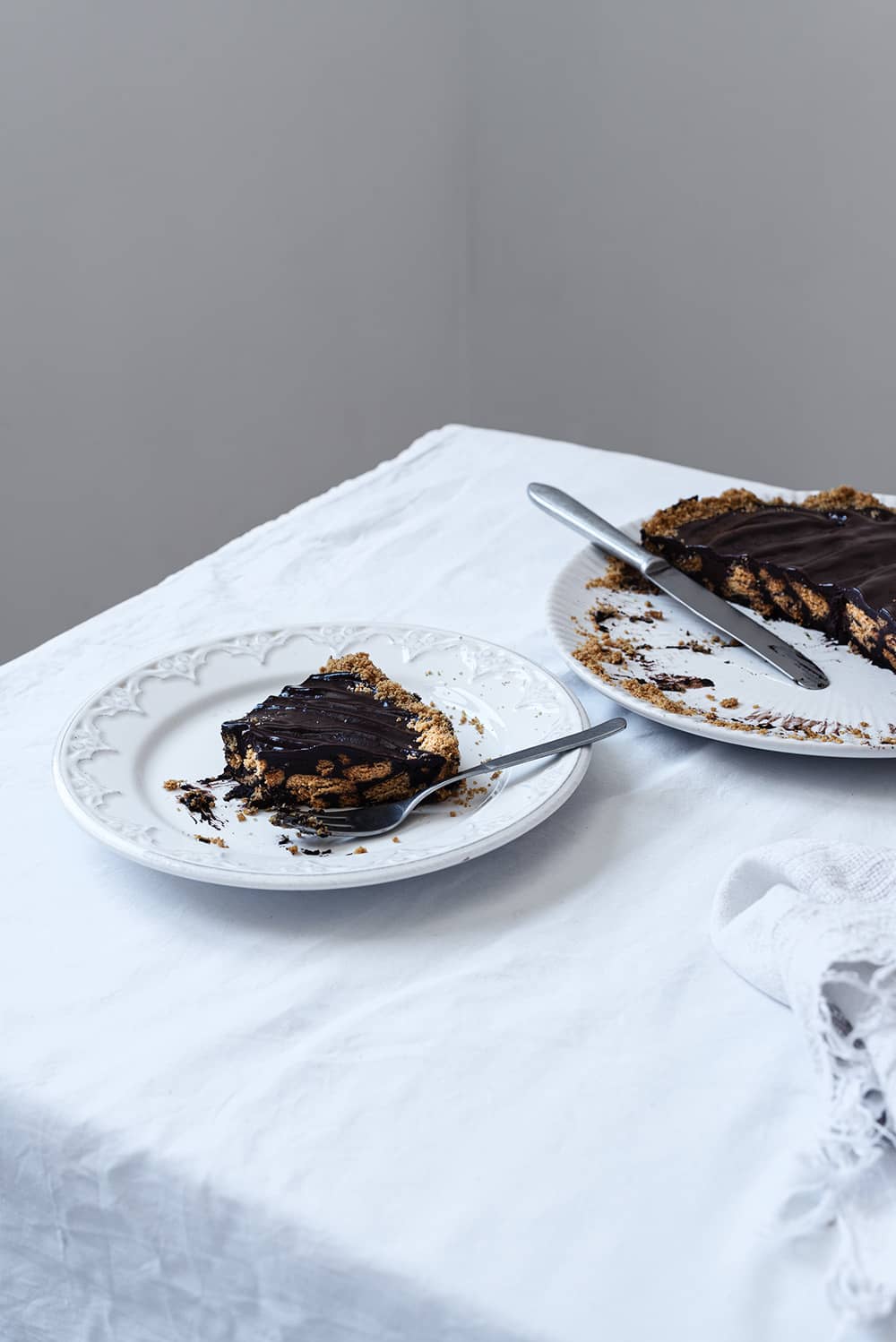 En una mesa cubierta con mantel blanco hay un plato blanco. Encima está la tarta de chocolate con galleta ya cortada. Delante de la tarta hay un plato blanco más pequeño con una ración de esta tarta. Al lado del postre hay un tenedor.