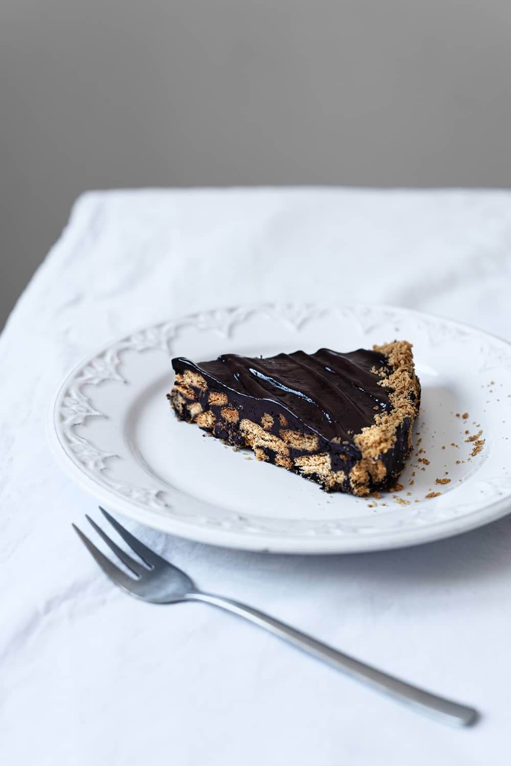 En una mesa cubierta con mantel blanco hay un plato blanco.En este plato hay una pequeña ración de la tarta de chocolate con galleta. Al lado del plato hay un tenedor. 