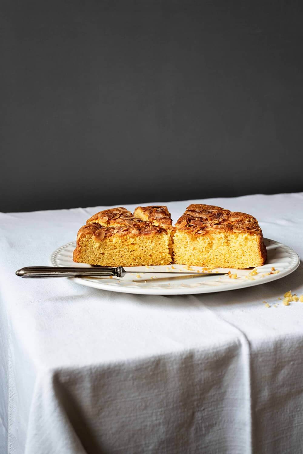 En una mesa, cubierta con mantel blanco hay un plato blanco. En el plato está el bizcocho de almendra de la abuela cortado. Se ve su miga tierna. En el plato hay un cuchillo también. 