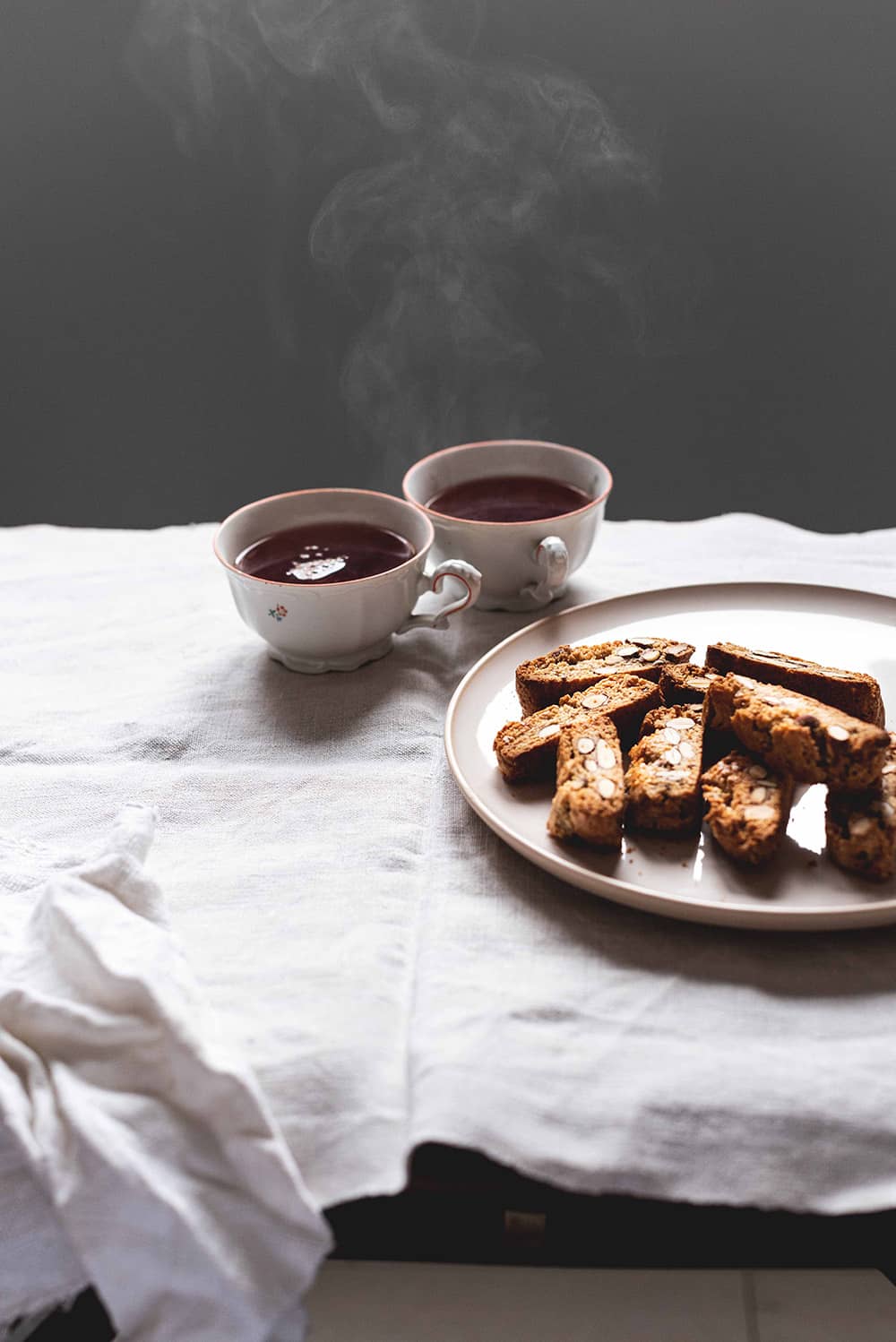 En una mesa hay un mantel blanco. En la mesa hay un plato blanco con Carquiñolis de almendra caseras. Al lado del plato hay dos tazas de té muy caliente y una servilleta de algodón.