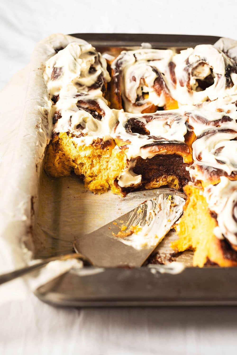 En una mesa, cubierta con mantel blanco, hay un molde rectangular. En el molde está el Cinnamon Rolls de calabaza. Ya está cortada. Se ve su textura esponjosa y miga tierna. En el molde está una pala para tartas.