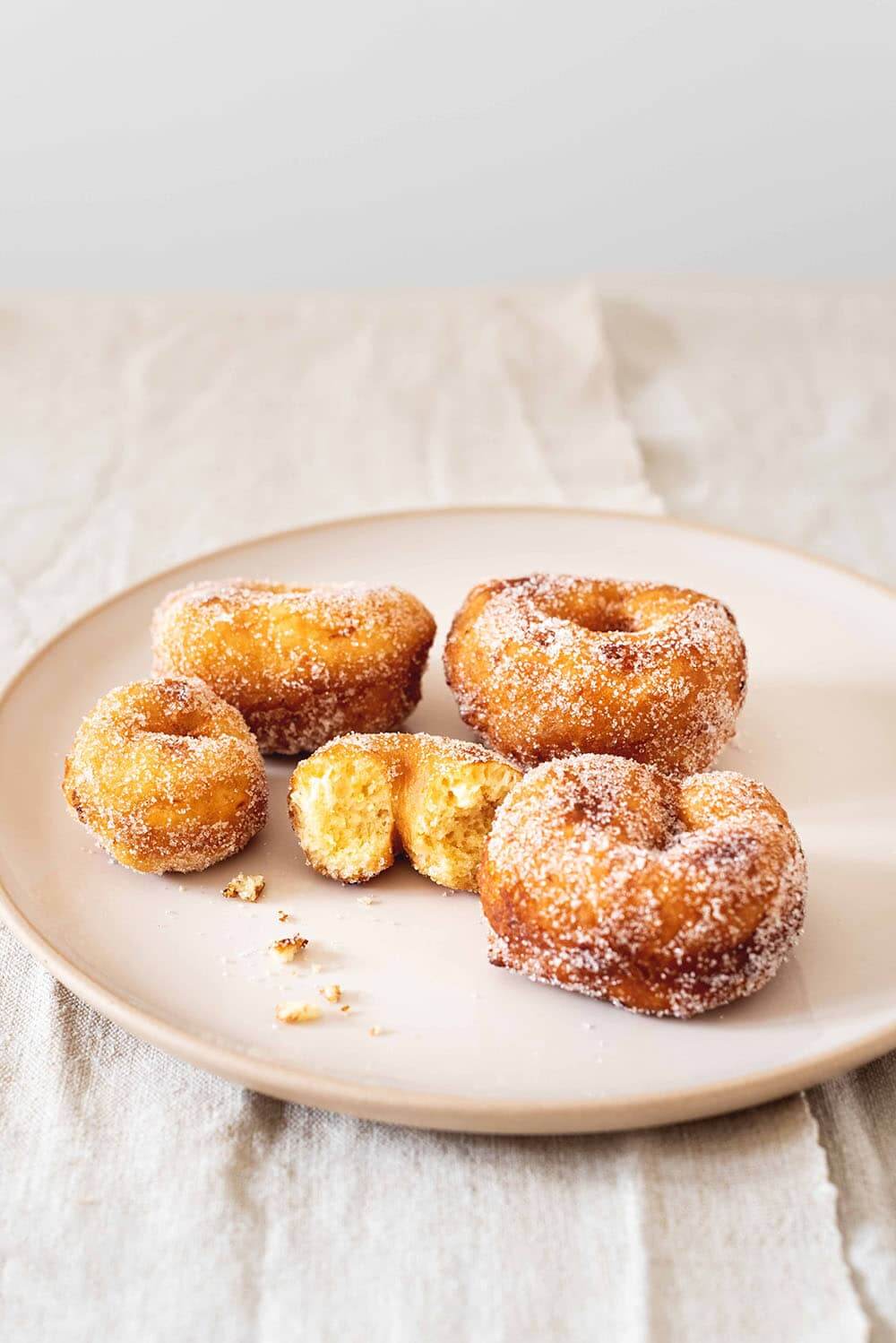 En una mesa hay un mantel blanco. En la mesa hay un plato blanco y en el plato hay cinco rosquillas de limón caseras y esponjosas. Una de ellas está mordida. Se ve tierna.