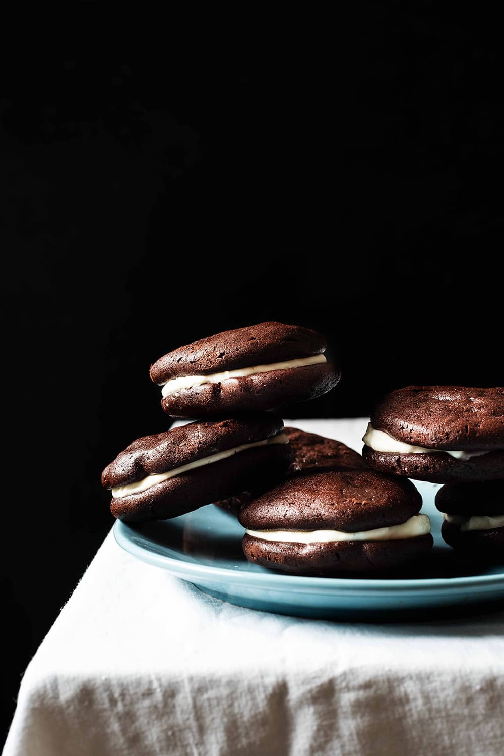 En una mesa hay un mantel blanco. En la mesa en un plato azul hay seis Whoopie pie de chocolate.