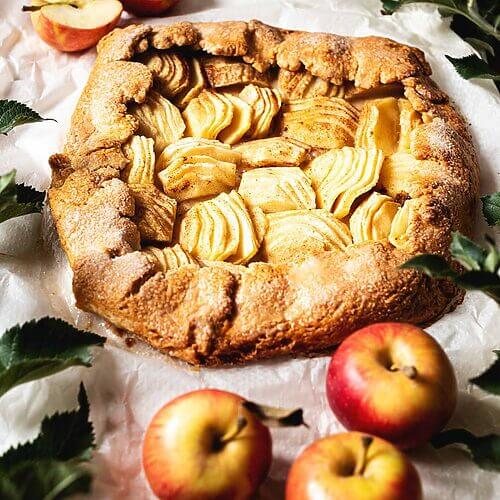 En una mesa de madera hay un papel de horno. En este papel está el galette de manzana y canela entera. Alrededor del postre hay varias manzanas y hojas verdes.