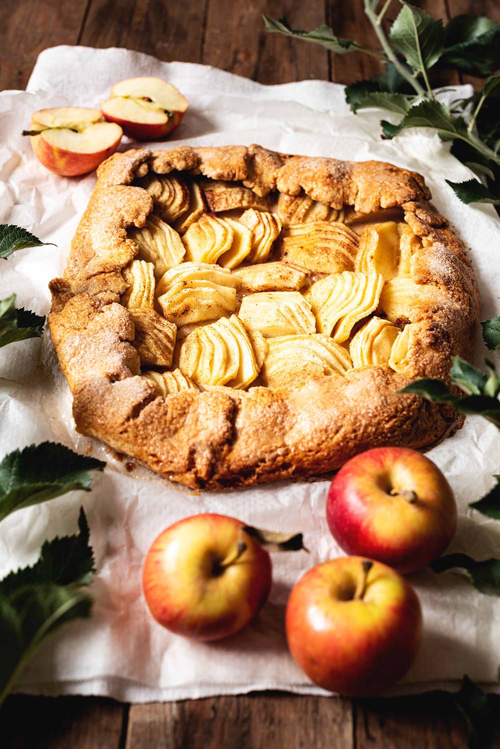 En una mesa de madera hay un papel de horno. En este papel está el galette de manzana y canela entera. Alrededor del postre hay varias manzanas y hojas verdes.