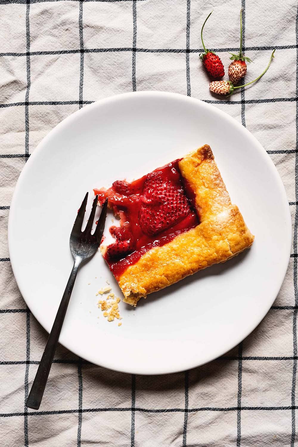 En la mesa hay un paño de cocina blanca con cuadros azules. En el paño hay un plato blanco. Encima hay un trozo de galette de fresas. Al lado de este trozo hay un tenedor.