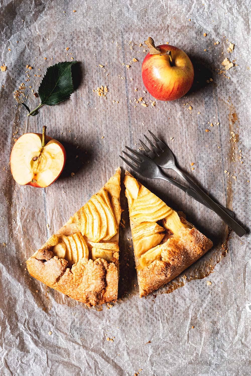 En una mesa de madera hay un papel de horno. En este papel hay dos trozos de galette de manzana y canela, dos tenedores, dos mitades de manzanas y una hoja verde. 