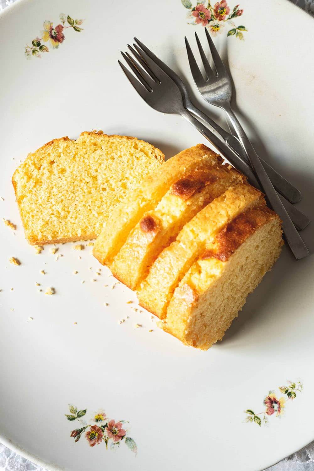 En una mesa hay un mantel blanco. En esta mesa se ve un plato grande blanco vintage. En el plato hay seis rodajas de Madera cake esponjoso. Al lado del bizcochos hay un par de tenedores.