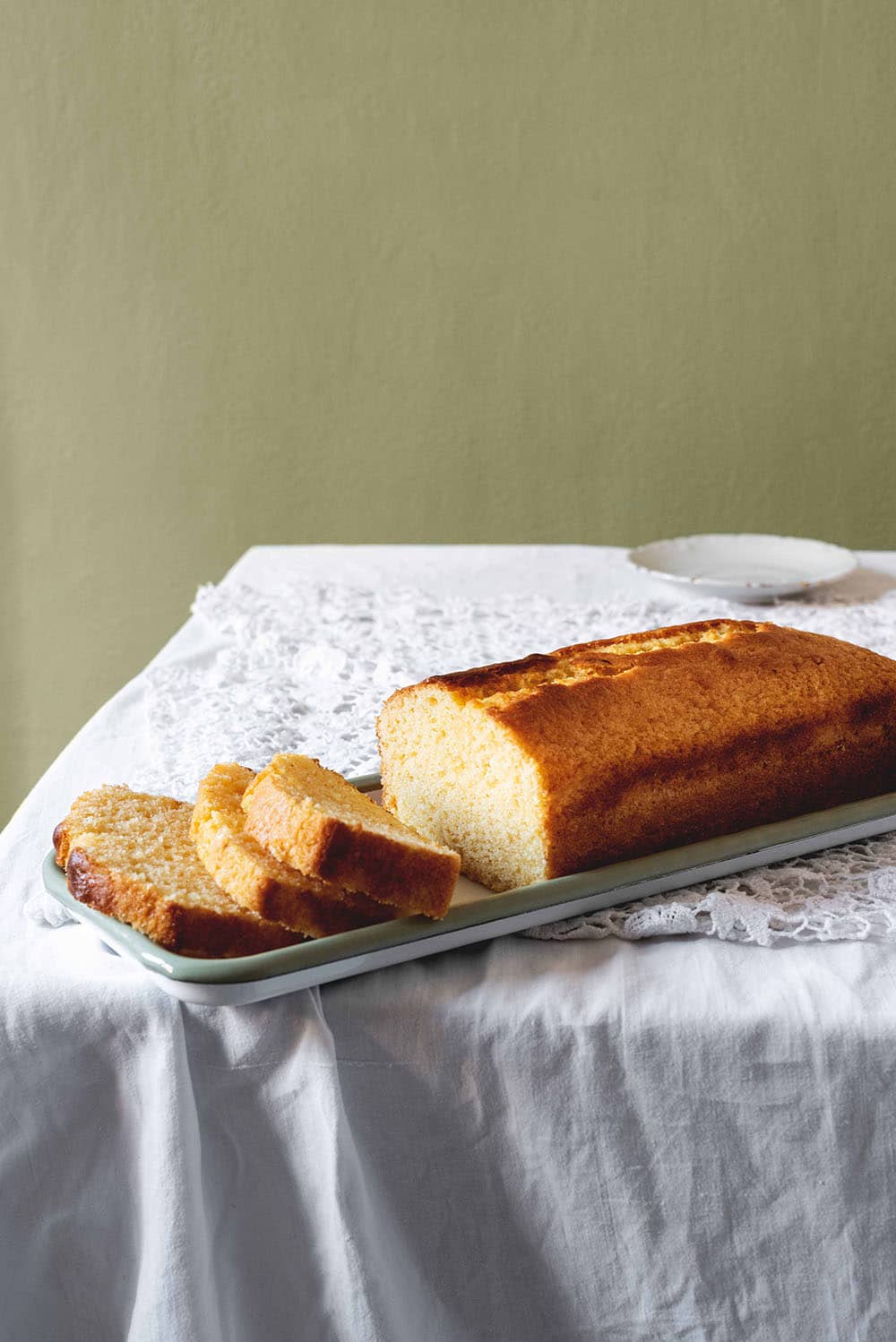 En una mesa hay un mantel blanco. En la mesa hay una bandeja blanca. En la bandeja está el Madeira cake.Ya está cortado la mitad del bizcocho.