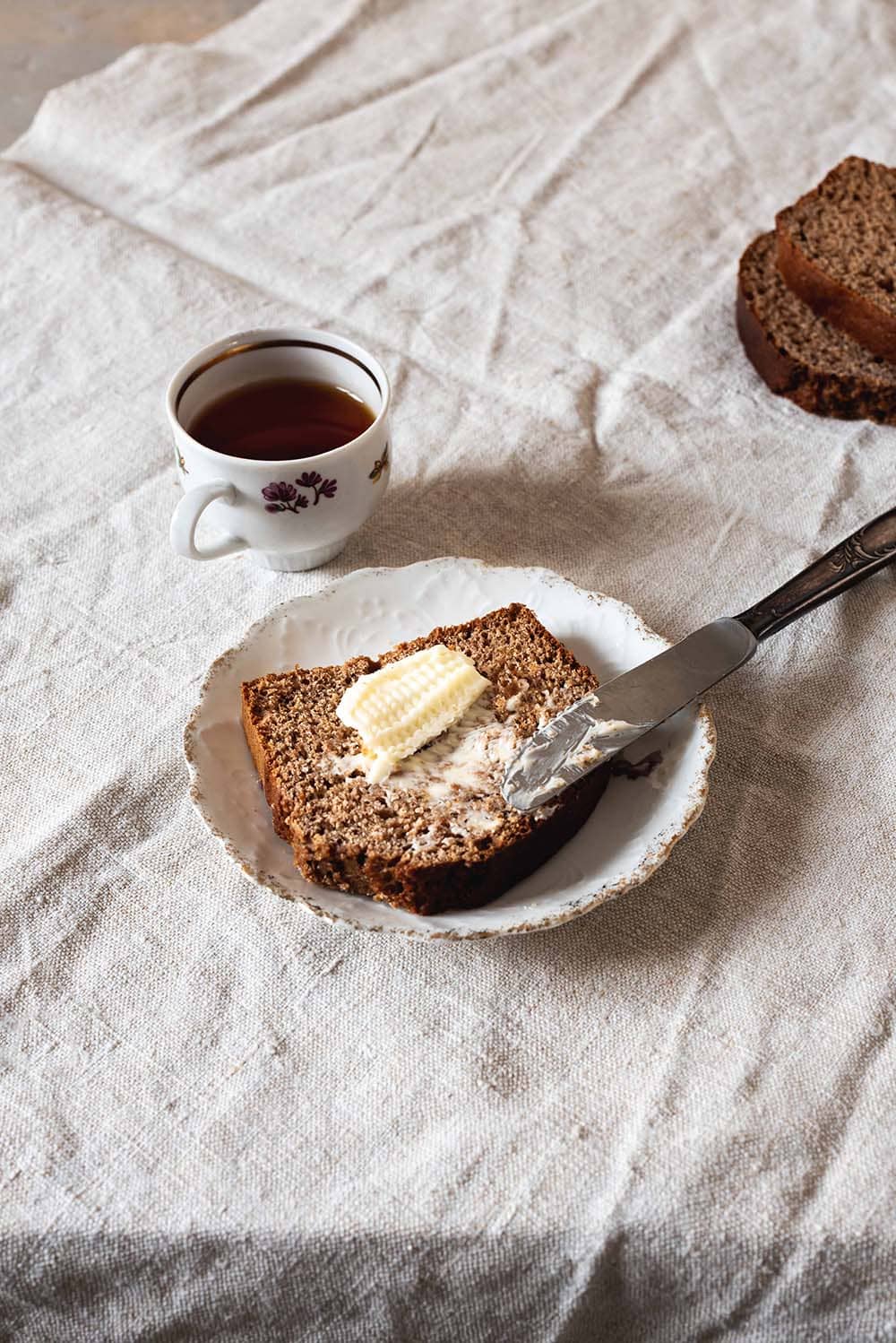 En una mesa cubierta con un mantel blanco hay un plato blanco. En el plato hay una rodaja de pan de especias. Encima hay un poco de mantequilla. Al lado del plato hay una taza de té.