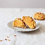En una mesa hay un mantel blanco. En un plato blanco vintage hay un Panellets de almendra con boniato cortado en la mitad. Detrás de este plato hay otro Panellets de almendra con boniato entero.