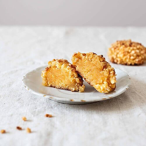 En una mesa hay un mantel blanco. En un plato blanco vintage hay un Panellets de almendra con boniato cortado en la mitad. Detrás de este plato hay otro Panellets de almendra con boniato entero.