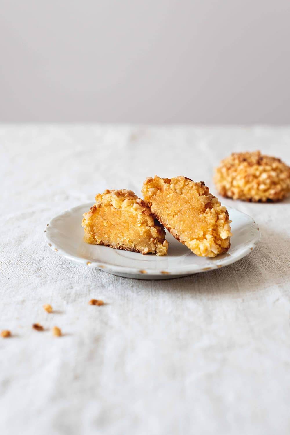 En una mesa hay un mantel blanco. En un plato blanco vintage hay un Panellets de almendra con boniato cortado en la mitad. Detrás de este plato hay otro Panellets de almendra con boniato entero.