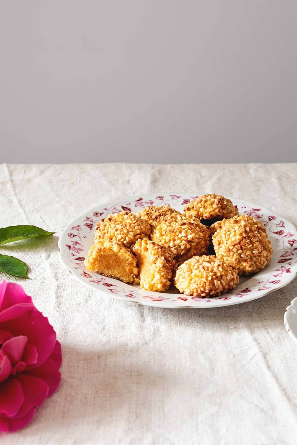 En una mesa hay un mantel blanco. En un plato blanco vintage hay muchos Panellets de almendra. Uno de ellos está cortado en la mitad. Se ve muy jugoso. Delante de este plato hay una rosa.